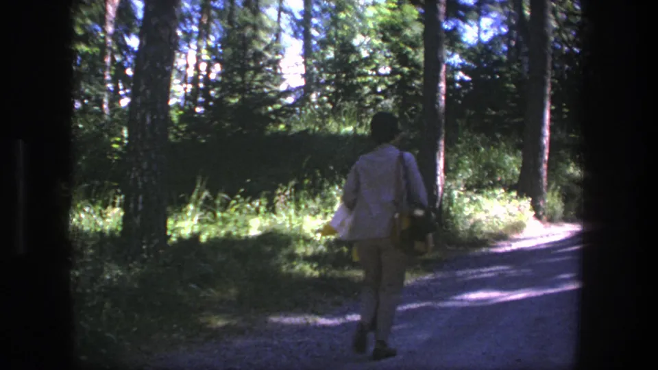 A person continues away from the camera on a wooded Danish dirt road, carrying a bag and yellow object.