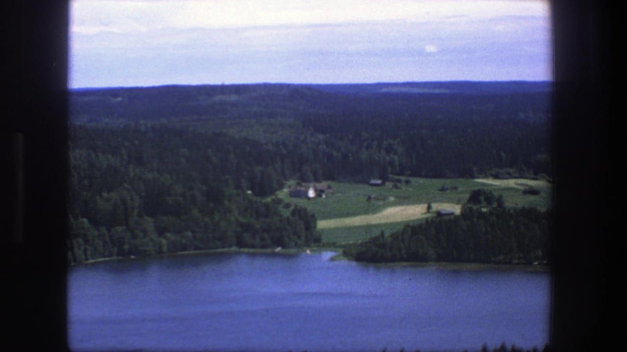 1975 Danish 8mm frame showing a lake below forested hills, green fields and small farm buildings from an elevated viewpoint.