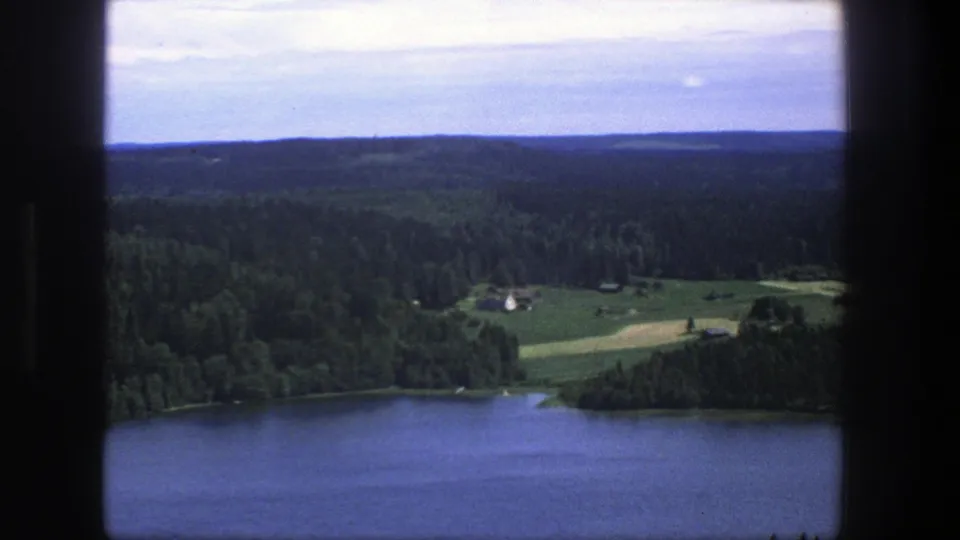 Danish 1975 home-movie frame of a broad lake, conifer forest, open fields and a small farm building under a pale sky.