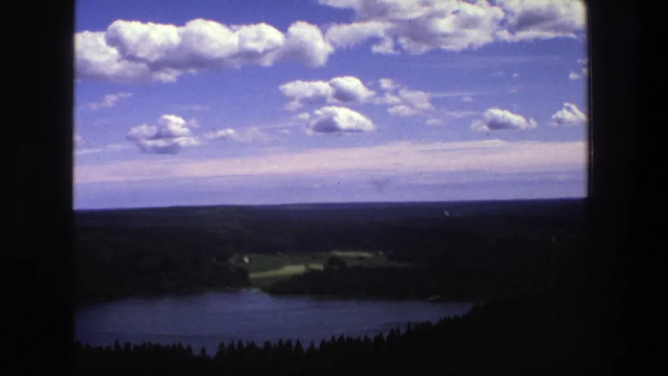 1975 Denmark 8mm frame with a darker exposed lake, silhouetted forest edge, fields, low horizon and scattered white clouds.
