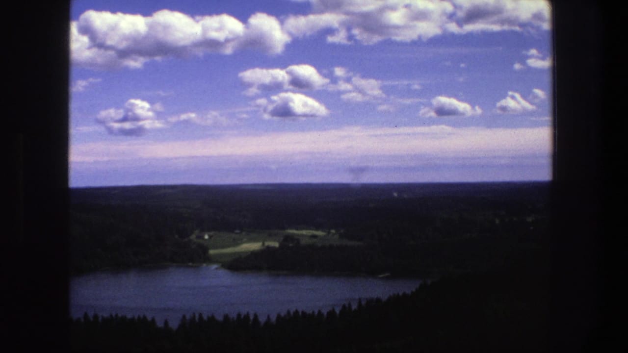Blue lake below dark forest with distant fields, horizon, clouds, and dark window edges.