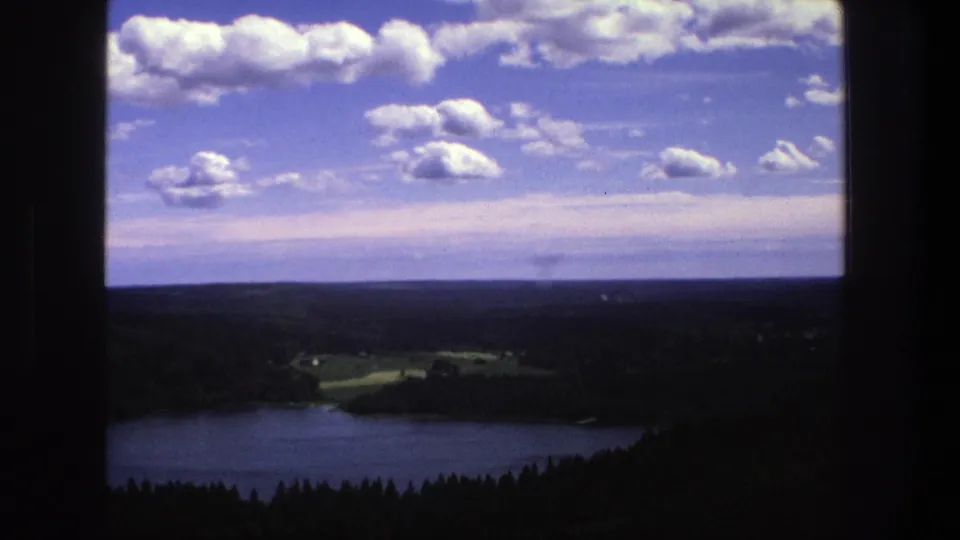 1975 Denmark 8mm view over a blue lake, forested shoreline, distant fields, and puffy clouds framed by dark window edges.