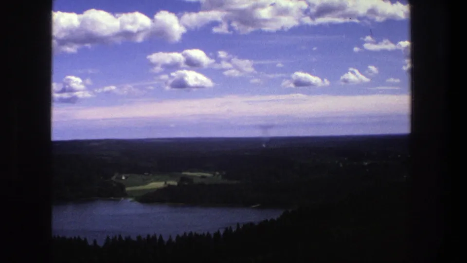 1975 Denmark home-movie frame showing a lake and wooded terrain under bright clouds, with the view bordered by dark window or film edges.