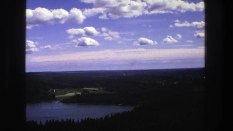 1975 Denmark 8mm landscape view with the lake partly visible below, dark woods in the foreground, fields beyond, and scattered clouds overhead.