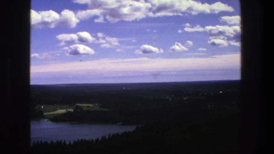 1975 Denmark home-movie view of a lake and wooded landscape under scattered white clouds, framed by dark vertical edges from the shooting position.