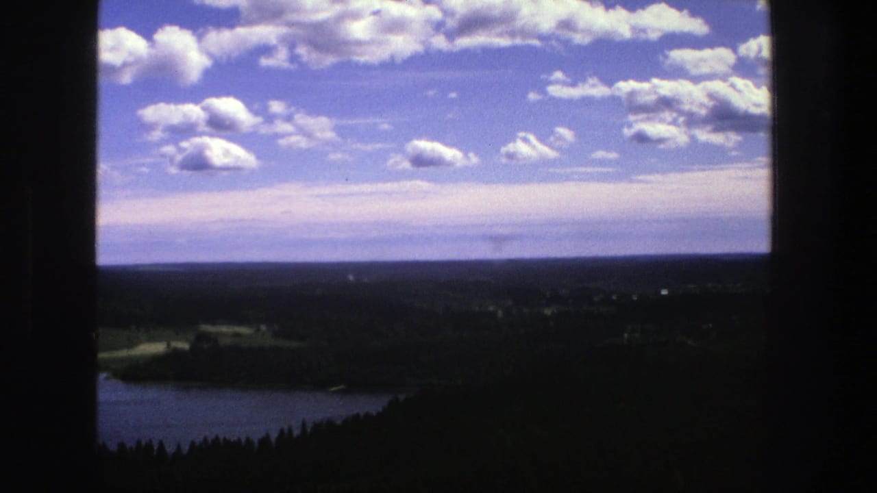 Hilltop view over lake, forest, fields, horizon, and cloudy blue sky framed by dark edges.