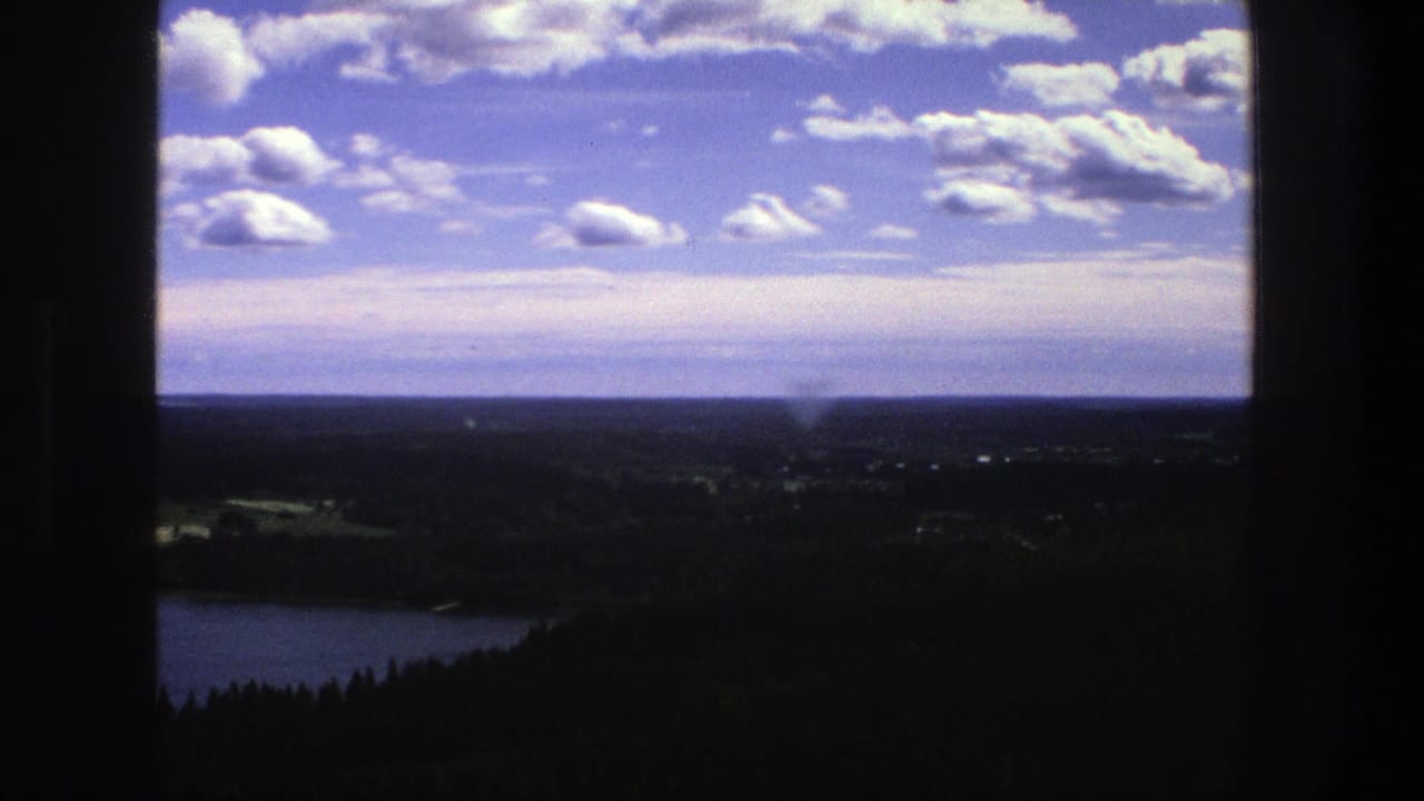Wide countryside view with lake, dark foreground, distant fields, horizon, and cloud-filled sky.