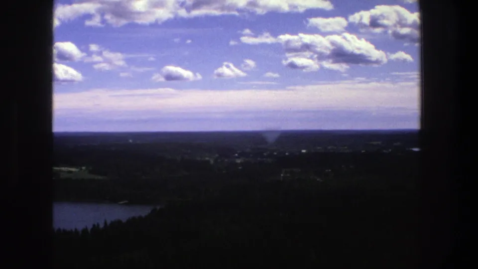 1975 Denmark 8mm frame showing a sweeping landscape with a lake edge, dark forest below, distant countryside, and scattered white clouds.