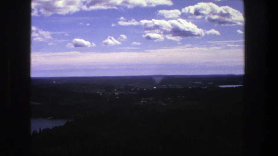1975 Denmark 8mm frame capturing a distant countryside panorama with lake, forested foreground, fields, horizon, and dark vertical framing.