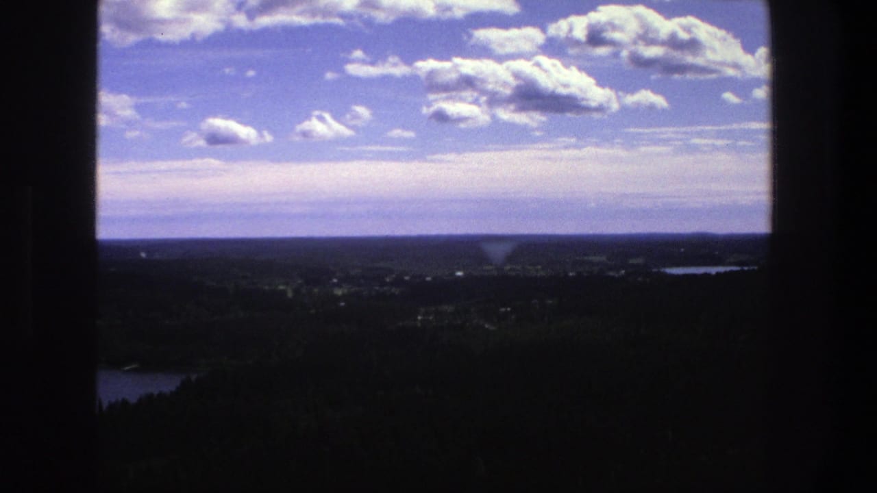 1975 Danish 8mm frame showing an elevated landscape view through dark window trim, with forest, distant water, horizon, and cumulus clouds.