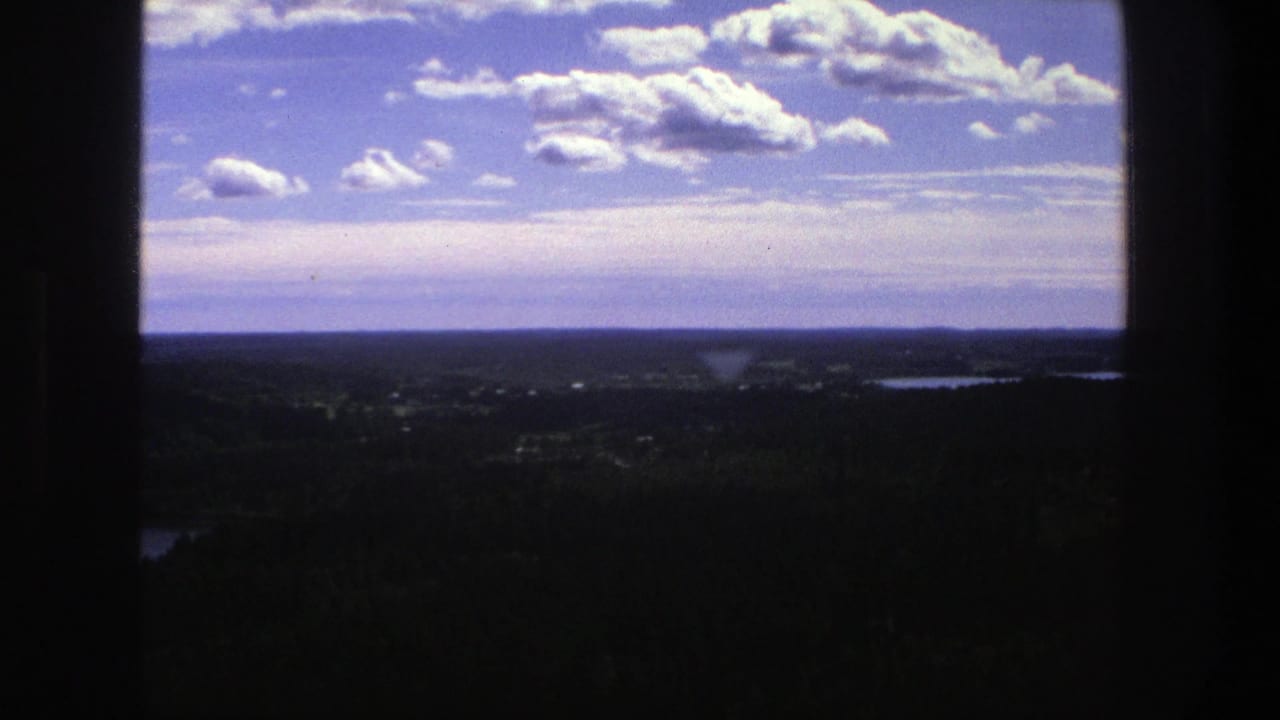 1975 Danish 8mm view across a dark foreground frame toward forested land, distant lakes, a low horizon, and bright cumulus clouds.