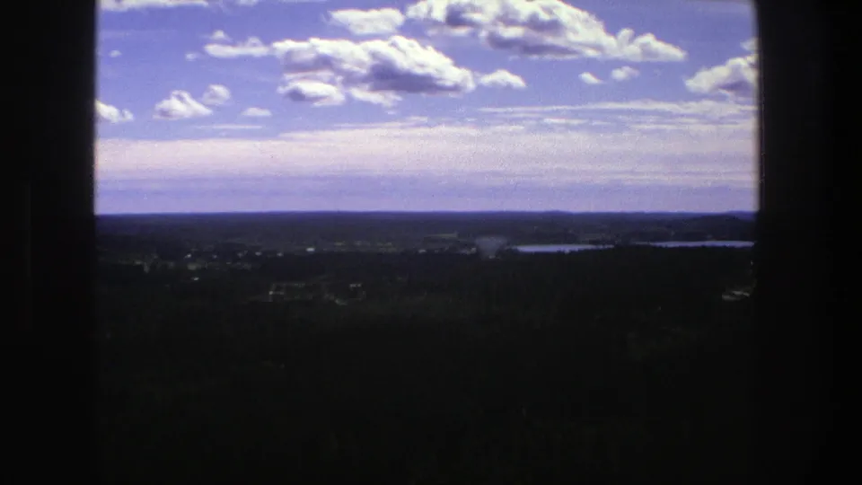 1975 Danish home-movie frame with a window-bordered overlook toward forest, lakes, distant clearings, and a bright sky with clouds.