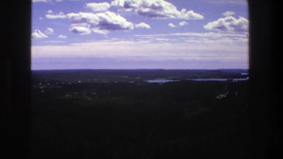 1975 Danish 8mm landscape frame showing dark foreground land, distant water, low horizon, and large cumulus clouds in a blue sky.