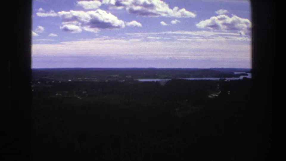 1975 Denmark 8mm view of a distant treeline and water beneath scattered clouds, partly obscured by dark foreground edges.