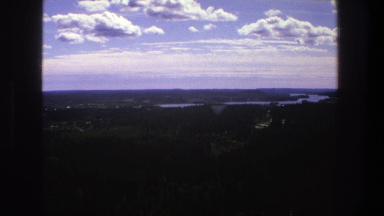 1975 Denmark 8mm frame showing a broad rural horizon, distant water, dark forest, and bright clouds from an elevated viewpoint.