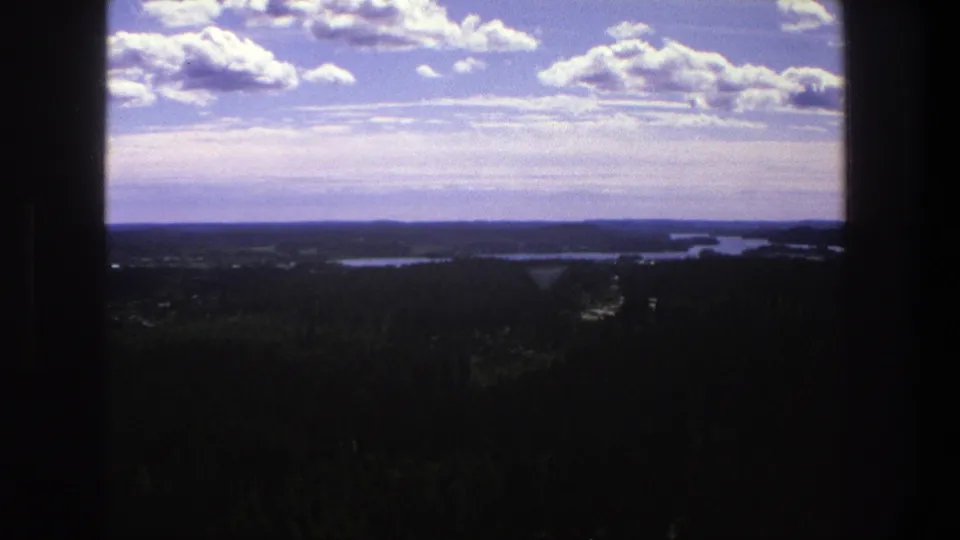 1975 Denmark 8mm landscape with blue sky, white clouds, dark trees, and distant water seen through shadowed frame edges.