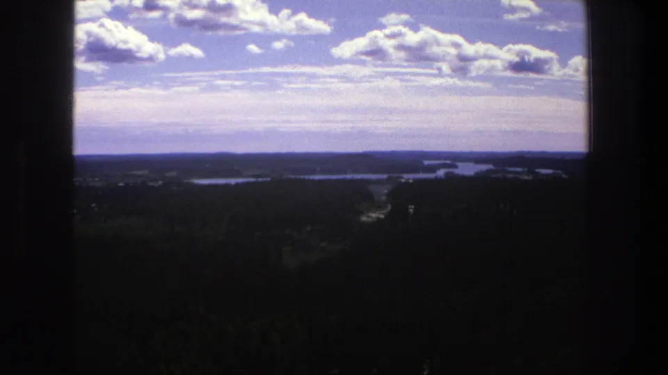 1975 Denmark 8mm view of a lake or river beyond dark forest, with scattered clouds across a pale blue sky.