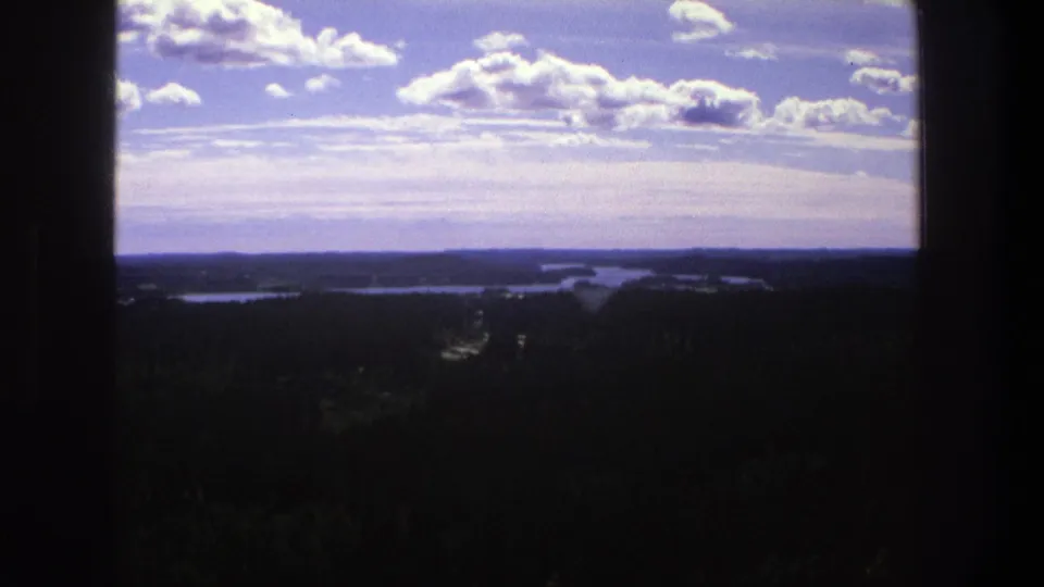 1975 Denmark home-movie frame with a high scenic view over dark woodland, reflective waterways, low hills, and scattered clouds.