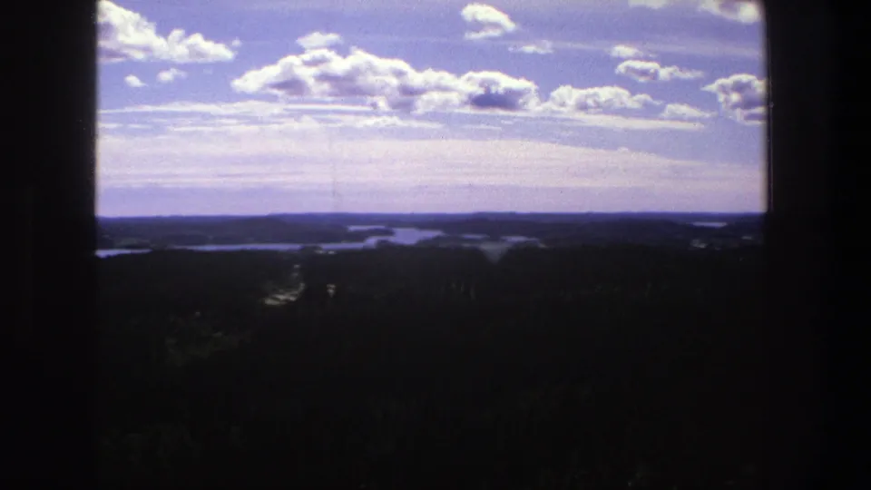 1975 Denmark 8mm landscape frame showing a high lookout view over wooded terrain, distant water, low horizon, and textured clouds.