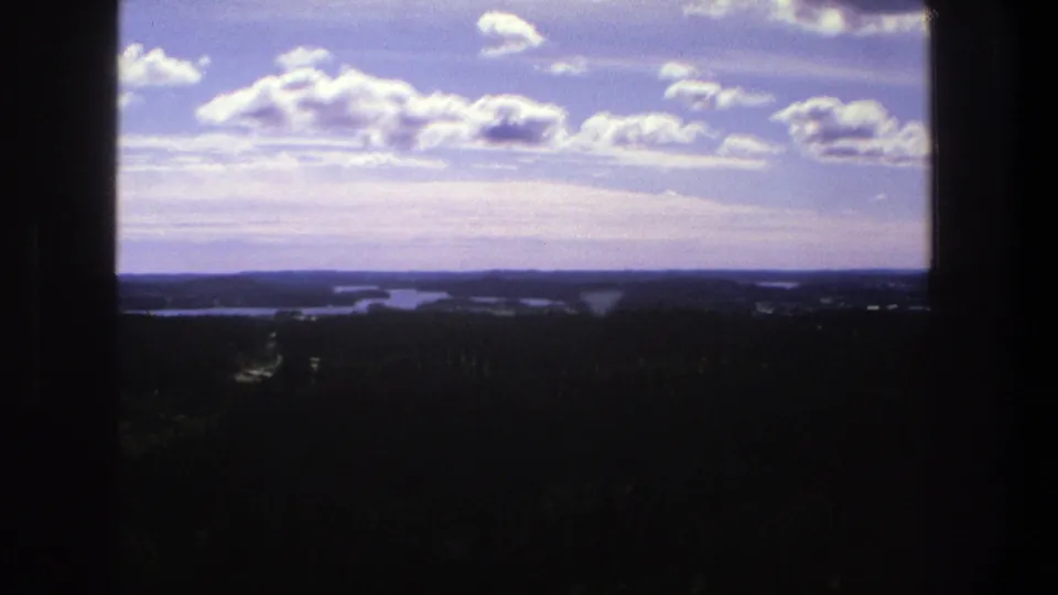 1975 Denmark home-movie frame of a distant rural panorama, with waterways and forest beneath a bright sky framed by dark edges.