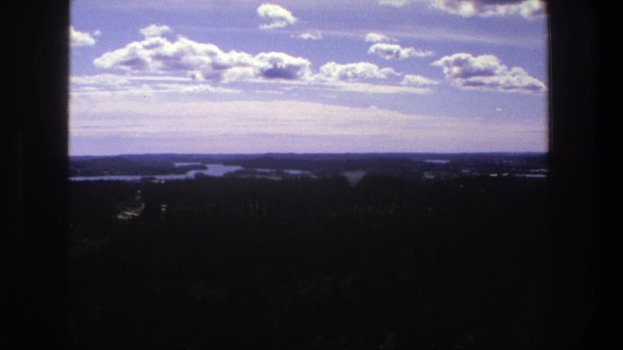 Blue sky with scattered clouds above a dark forested horizon and distant lakes.