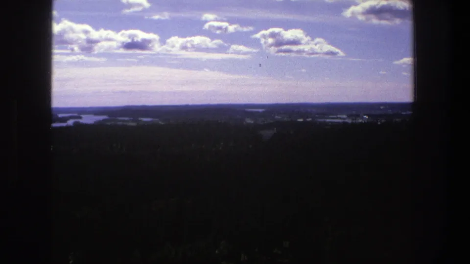 1975 Denmark 8mm view of a clouded blue sky over dark forest, with small lake forms along the far horizon.