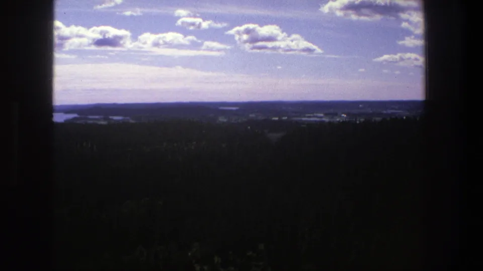 1975 Denmark 8mm frame showing a wide sky with bright clouds over a shadowed forest and lake-dotted horizon.