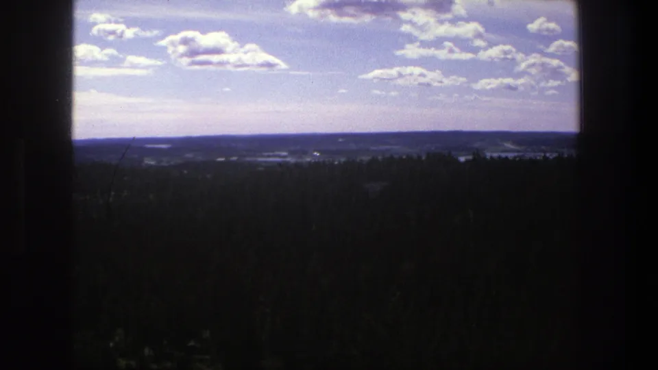 1975 Denmark 8mm frame showing a wide landscape with dark trees in the foreground, distant hills, and bright summer clouds.