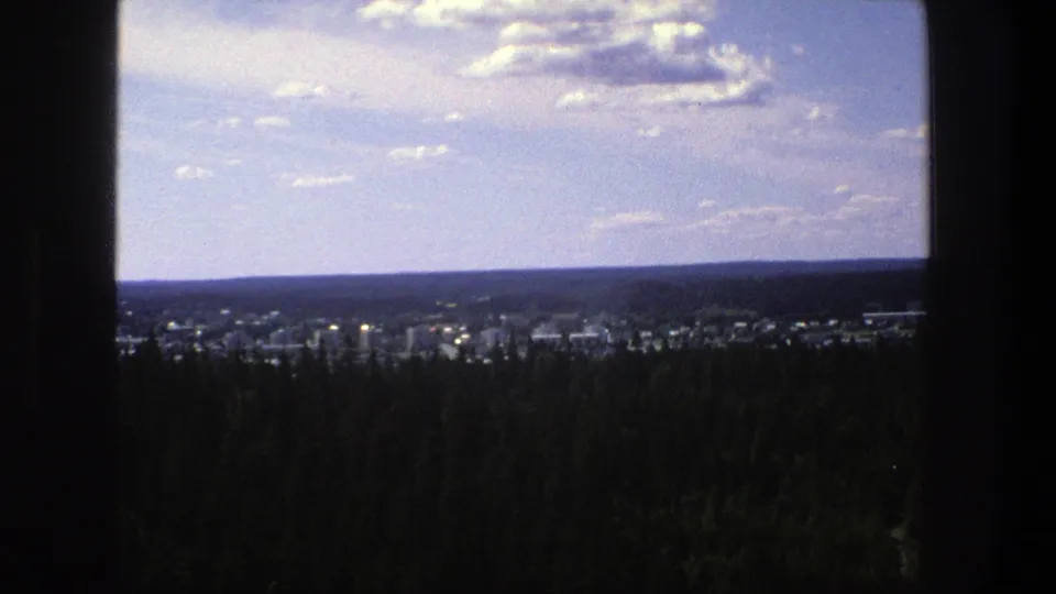 1975 Denmark 8mm frame showing a distant town, dark pine forest foreground, and scattered clouds across the sky.