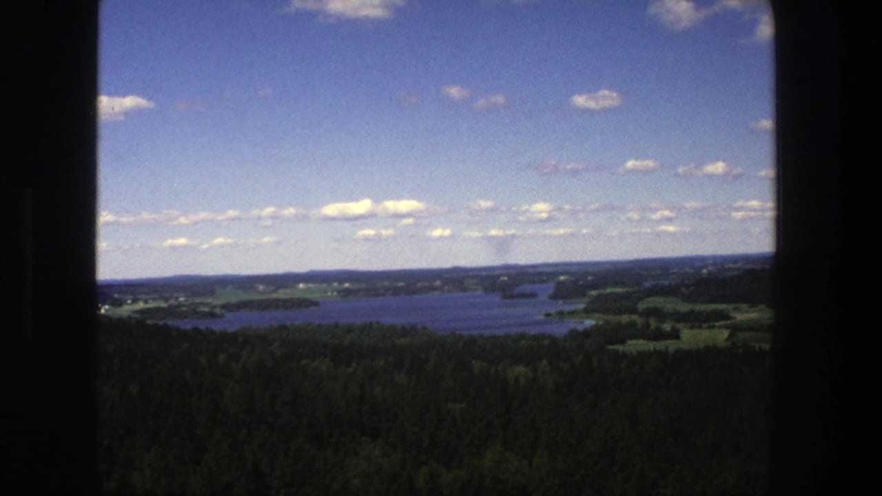 Elevated view of a lake, forest, fields, and blue sky framed by dark film edges.