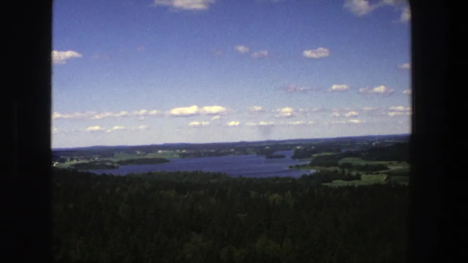 1975 Denmark 8mm frame showing a high view over lakes, woodland, distant fields, and a cloud-dotted blue sky.