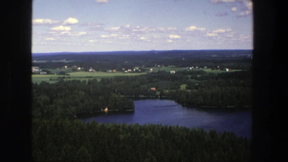1975 Denmark 8mm frame with a wooded lakeshore, distant rural fields, small buildings, and a bright clouded sky.