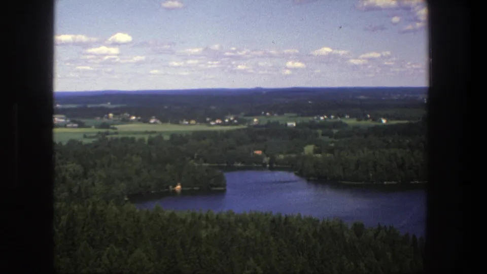 1975 Denmark 8mm frame looking over a dark blue lake, surrounding woods, rural fields, and distant settlement.