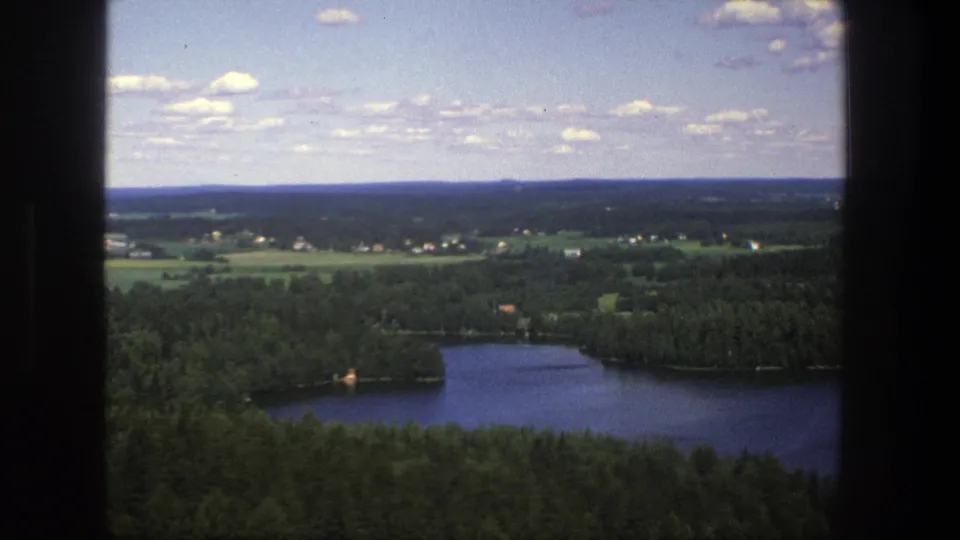1975 Denmark 8mm frame showing a lake bordered by forest with fields, distant houses, and pale clouds overhead.