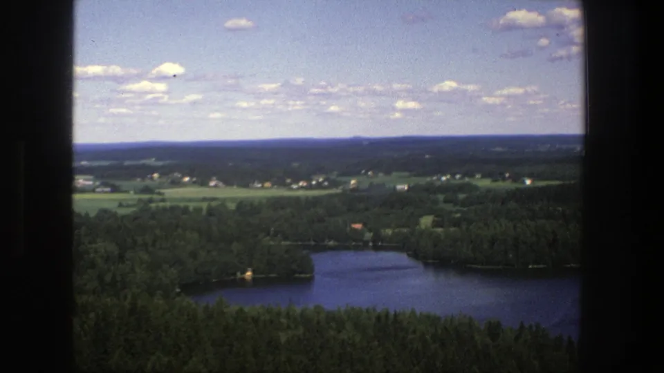 1975 8mm aerial frame over rural Denmark with a lake at the lower edge, forest, fields, small buildings, and soft film grain.