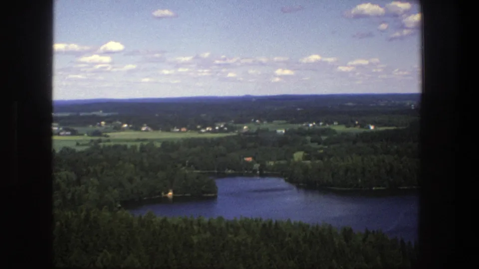Late-frame 1975 Denmark aerial view showing a wooded lake, rural fields, scattered homes, distant low hills, and partly cloudy sky.