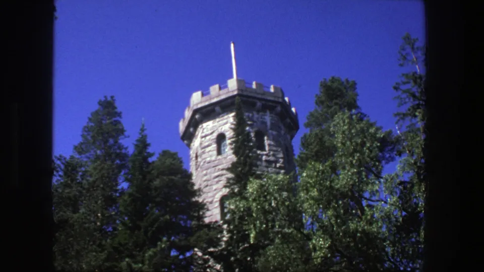Low-angle 1975 Denmark 8mm view of a stone tower among green trees, with its flagpole and upper wall visible.