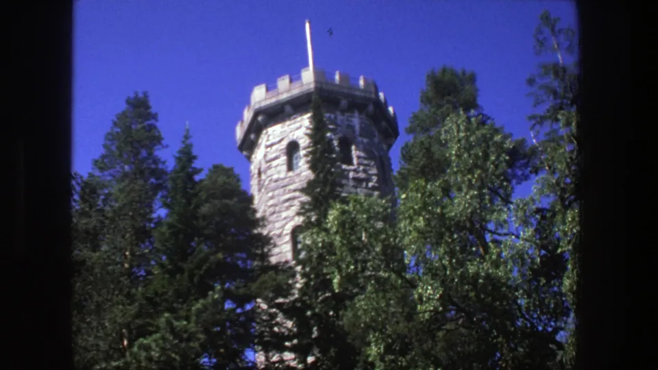 1975 Denmark 8mm frame of the stone tower above surrounding trees, with crenellations and flagpole visible in bright daylight.