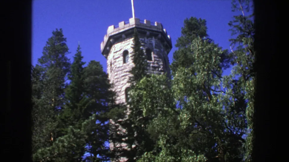 1975 Denmark 8mm frame showing a castle-like stone tower partly hidden by evergreen trees and bright blue sky.