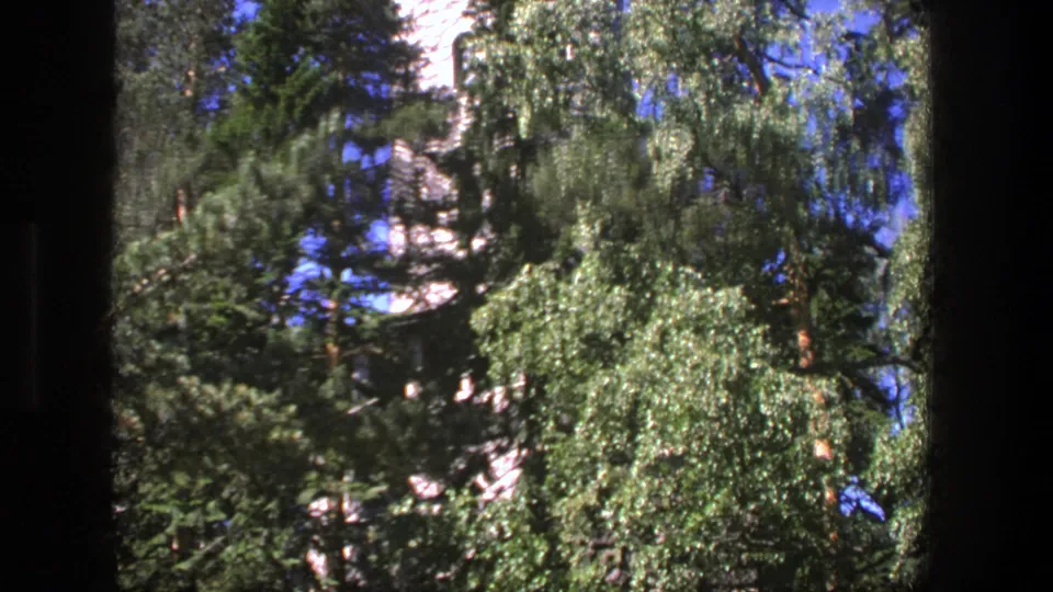 1975 Denmark 8mm frame of a castle-like tower mostly screened by evergreen and leafy trees beneath a clear sky.