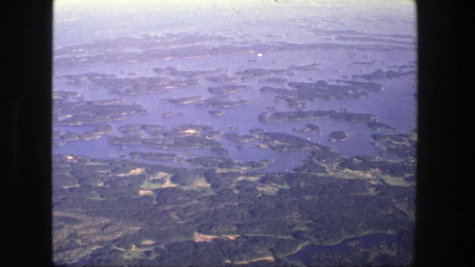1975 Denmark aerial home-movie frame looking over an archipelago-like landscape of islands, water, and dark wooded areas.