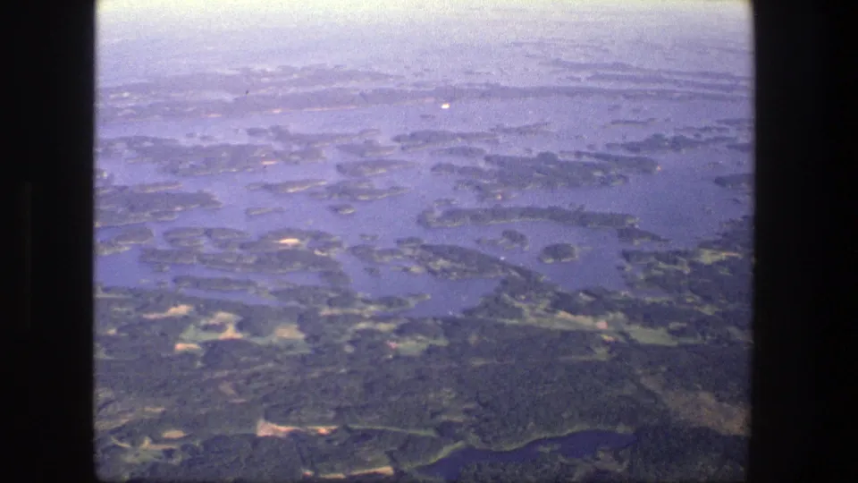 1975 Denmark 8mm aerial view over green wooded islands, blue water channels, and hazy distant shoreline.