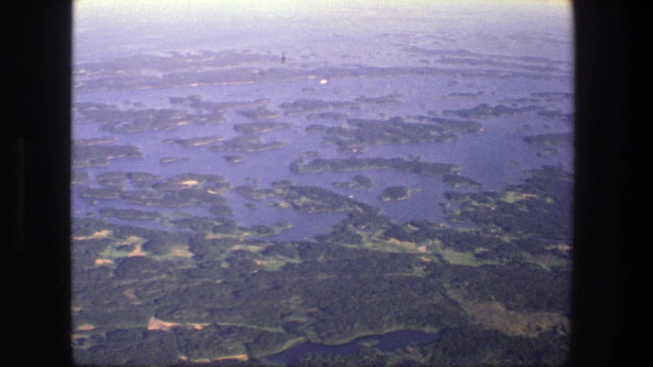 1975 Denmark aerial home-movie frame showing scattered wooded islands, blue waterways, and a pale hazy sky near the horizon.