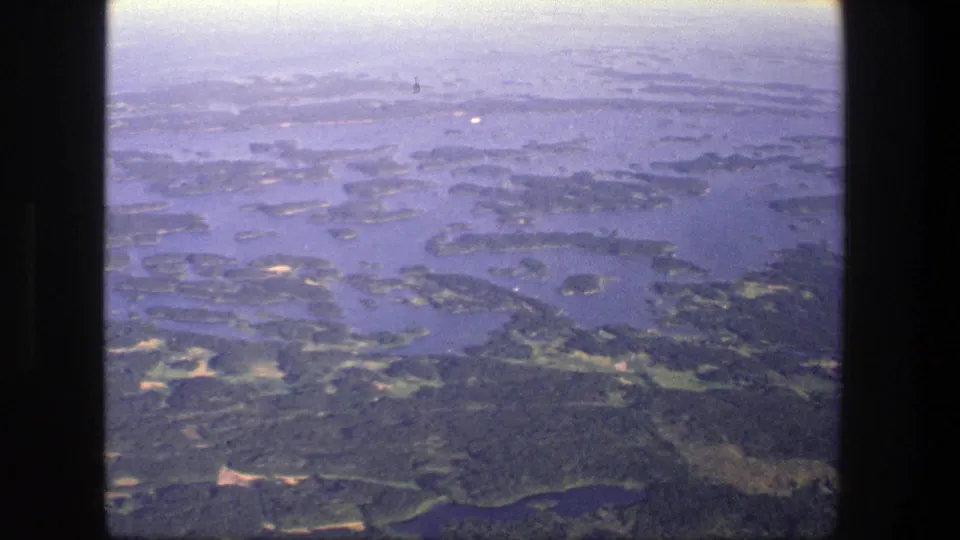 1975 Denmark aerial home-movie frame showing scattered wooded islands, blue waterways, and a pale hazy sky near the horizon.