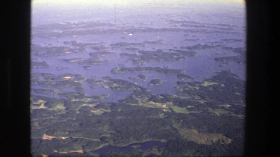 1975 Denmark 8mm frame with wooded land masses, winding blue water, small pale clearings, and soft aerial haze.