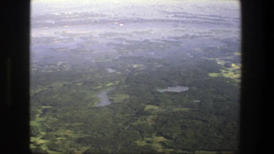 1975 Denmark 8mm aerial view over dense green woodland, small lakes, and a hazy horizon seen through dark frame edges.