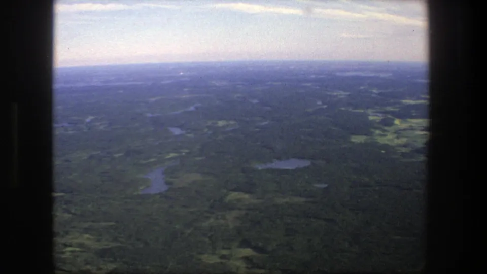 A 1975 Danish countryside aerial shot shows wooded terrain, reflective lakes, and a soft blue horizon from an airplane-window perspective.