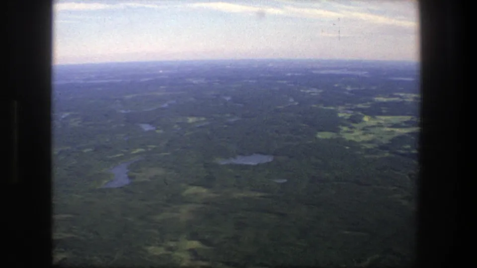 1975 Denmark 8mm frame of forested countryside and water bodies, with a dark central border and hazy sky emphasizing the home-movie scan.