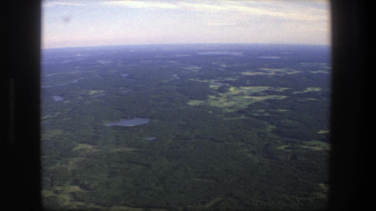 1975 Denmark 8mm aerial view over forest, scattered small lakes, open fields, and a pale hazy sky framed by dark film edges.