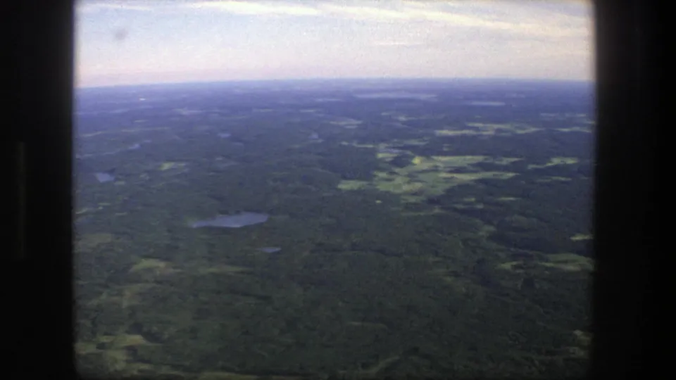 1975 Denmark 8mm aerial view over forest, scattered small lakes, open fields, and a pale hazy sky framed by dark film edges.